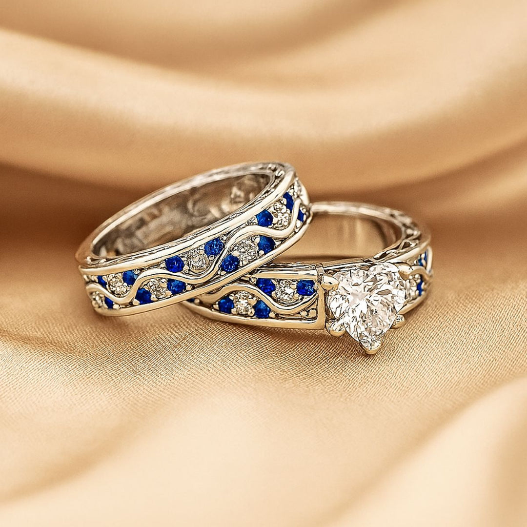 Fiesta ring set by CouplesChoices.com, featuring a radiant heart-cut diamond centerpiece with blue and white stones in a flowing wave design, paired with a silver and sapphire-toned textured band.