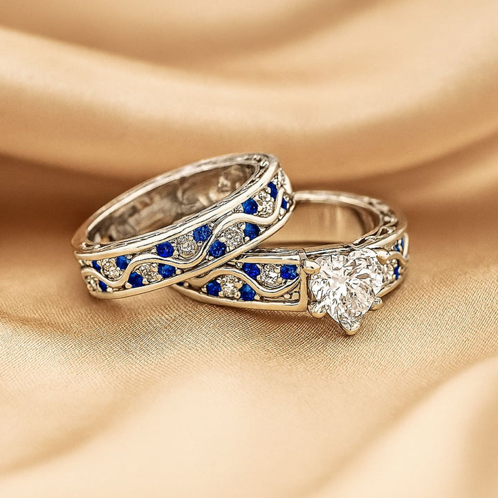 Fiesta ring set by CouplesChoices.com, featuring a radiant heart-cut diamond centerpiece with blue and white stones in a flowing wave design, paired with a silver and sapphire-toned textured band.