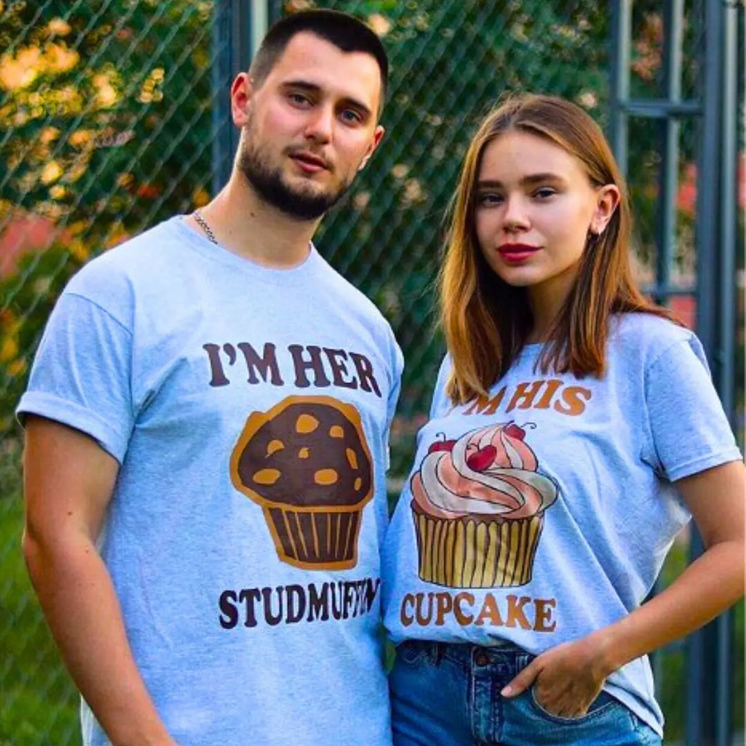 Adorably cheeky couple shirts in heather grey—hers says 'I’m His Cupcake' with a frosted treat, his reads 'I’m Her Studmuffin' with a chocolate muffin. A sweet and playful pair from CouplesChoices.com.