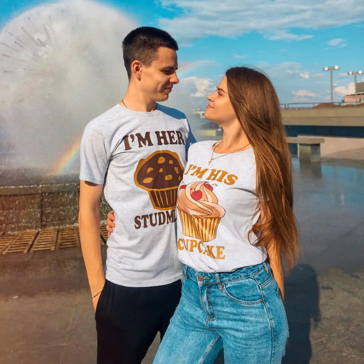 Adorably cheeky couple shirts in heather grey—hers says 'I’m His Cupcake' with a frosted treat, his reads 'I’m Her Studmuffin' with a chocolate muffin. A sweet and playful pair from CouplesChoices.com.