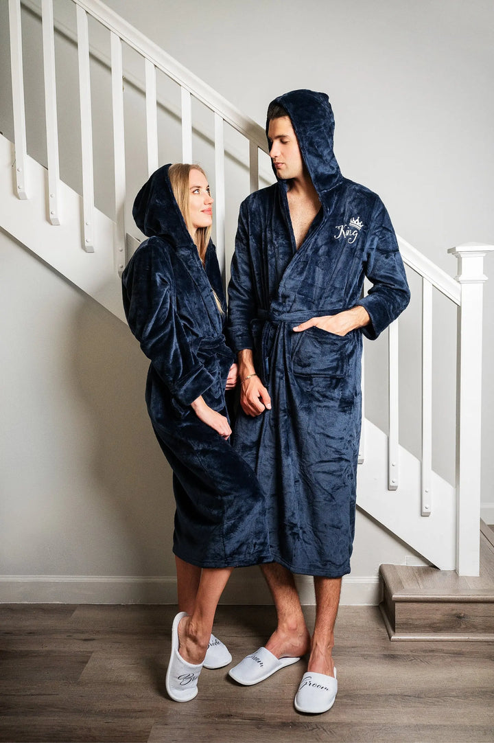 Couple wearing matching plush navy blue hooded bathrobes embroidered with elegant white “King” and “Queen” lettering and crown motifs—luxurious, cozy loungewear designed by CouplesChoices.com for modern royalty.