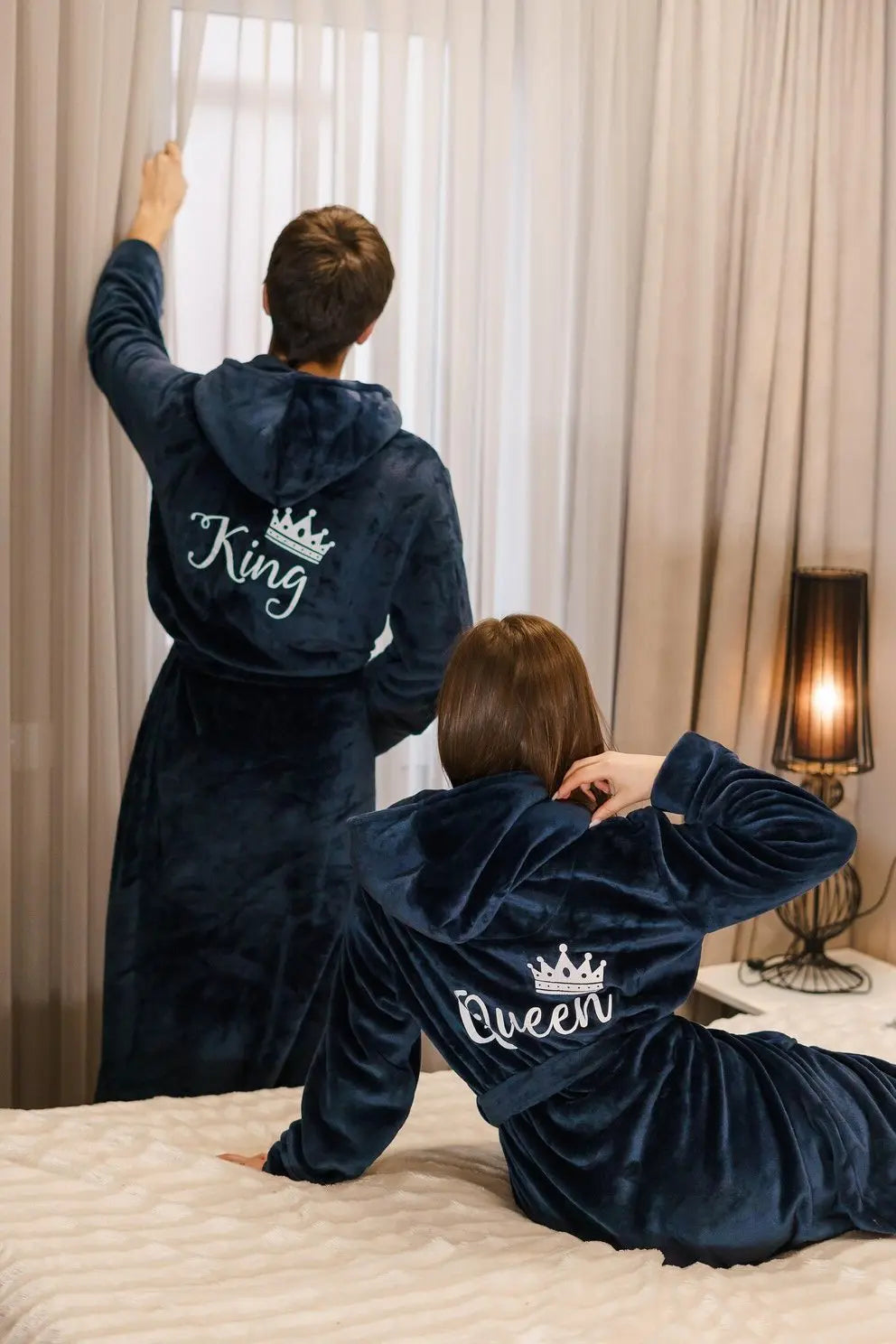 Couple wearing matching plush navy blue hooded bathrobes embroidered with elegant white “King” and “Queen” lettering and crown motifs—luxurious, cozy loungewear designed by CouplesChoices.com for modern royalty.