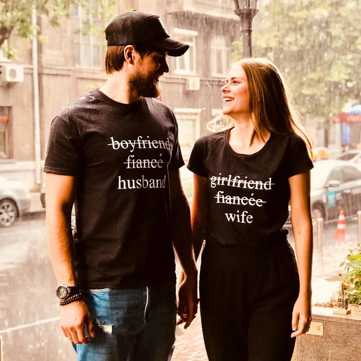 Couples' black matching tees—his shows 'boyfriend' and 'fiancé' crossed out with 'husband' below, hers reads 'girlfriend' and 'fiancée' crossed out with 'wife' below. A love story told in text, from CouplesChoices.com.