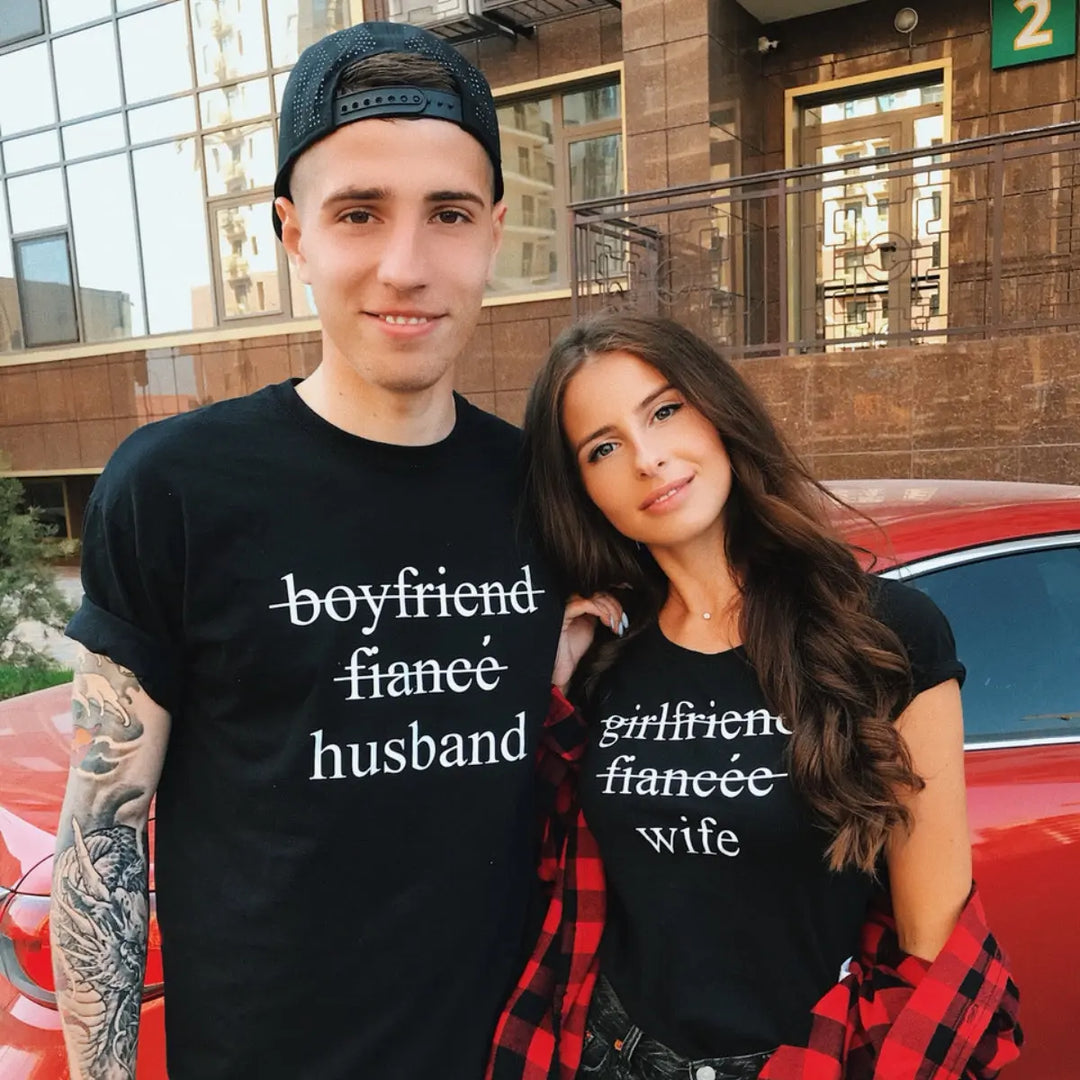 Couples' black matching tees—his shows 'boyfriend' and 'fiancé' crossed out with 'husband' below, hers reads 'girlfriend' and 'fiancée' crossed out with 'wife' below. A love story told in text, from CouplesChoices.com.