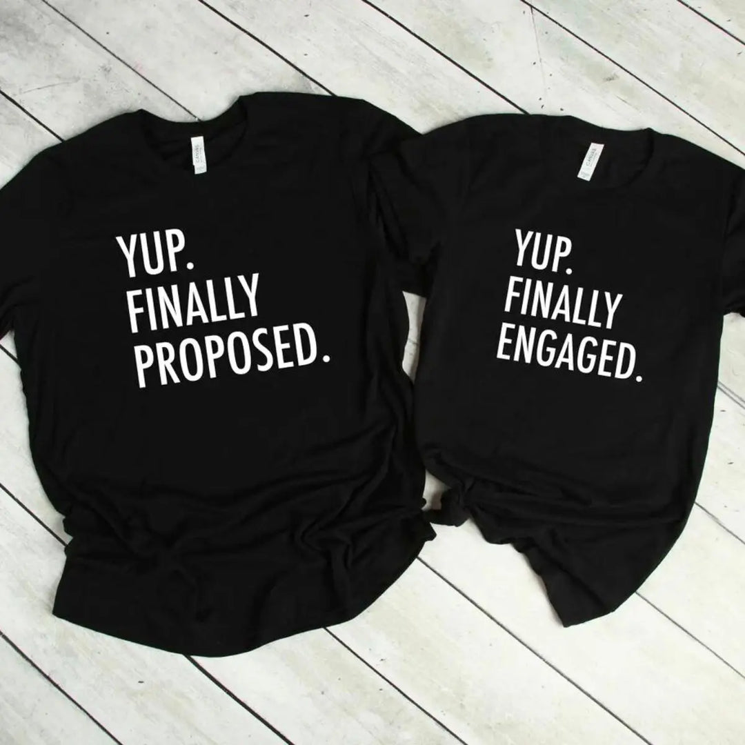 Couples' matching black tees—his says 'Yup. Finally Proposed.' and hers reads 'Yup. Finally Engaged.' Bold white text for couples ready to celebrate the moment. Exclusively from CouplesChoices.com.