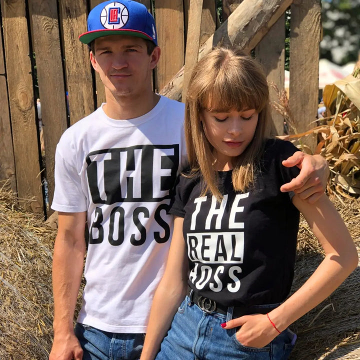 Couples matching graphic tees from CouplesChoices.com—his reads “THE BOSS” in bold black on white, hers says “THE REAL BOSS” in bold white on black. A playful and confident way to show off relationship dynamics with style.