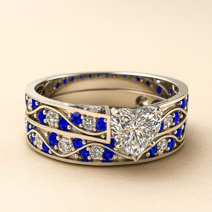 Fiesta ring set by CouplesChoices.com, featuring a radiant heart-cut diamond centerpiece with blue and white stones in a flowing wave design, paired with a silver and sapphire-toned textured band.