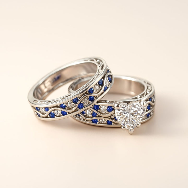 Fiesta ring set by CouplesChoices.com, featuring a radiant heart-cut diamond centerpiece with blue and white stones in a flowing wave design, paired with a silver and sapphire-toned textured band.