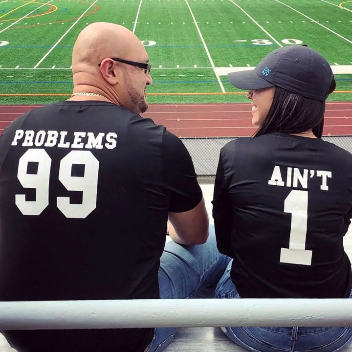 Matching black couple shirts—his reads '99 Problems' and hers answers 'Ain’t 1' in varsity-style white print. A playful nod to love and loyalty, exclusively from CouplesChoices.com.