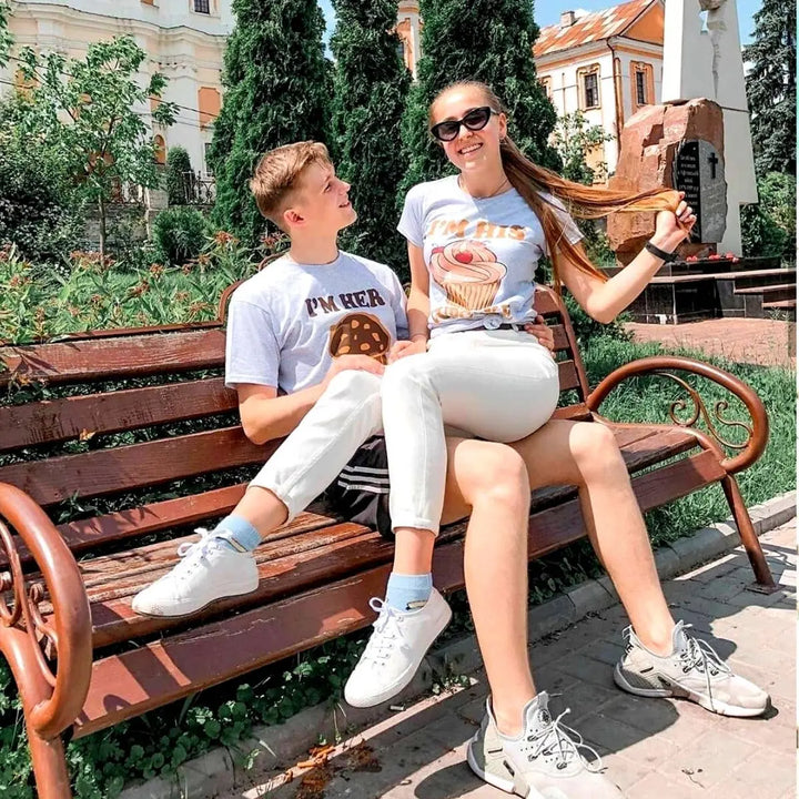 Adorably cheeky couple shirts in heather grey—hers says 'I’m His Cupcake' with a frosted treat, his reads 'I’m Her Studmuffin' with a chocolate muffin. A sweet and playful pair from CouplesChoices.com.