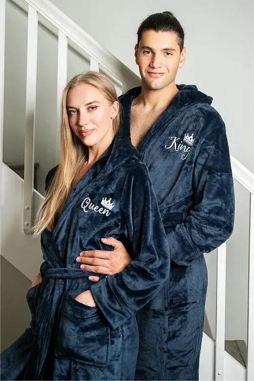Couple wearing matching plush navy blue hooded bathrobes embroidered with elegant white “King” and “Queen” lettering and crown motifs—luxurious, cozy loungewear designed by CouplesChoices.com for modern royalty.