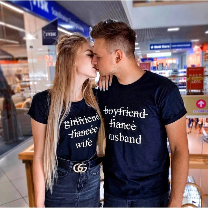 Couples' black matching tees—his shows 'boyfriend' and 'fiancé' crossed out with 'husband' below, hers reads 'girlfriend' and 'fiancée' crossed out with 'wife' below. A love story told in text, from CouplesChoices.com.