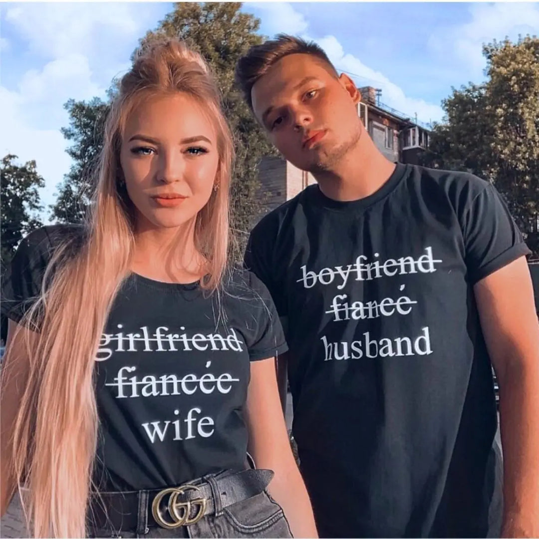 Couples' black matching tees—his shows 'boyfriend' and 'fiancé' crossed out with 'husband' below, hers reads 'girlfriend' and 'fiancée' crossed out with 'wife' below. A love story told in text, from CouplesChoices.com.