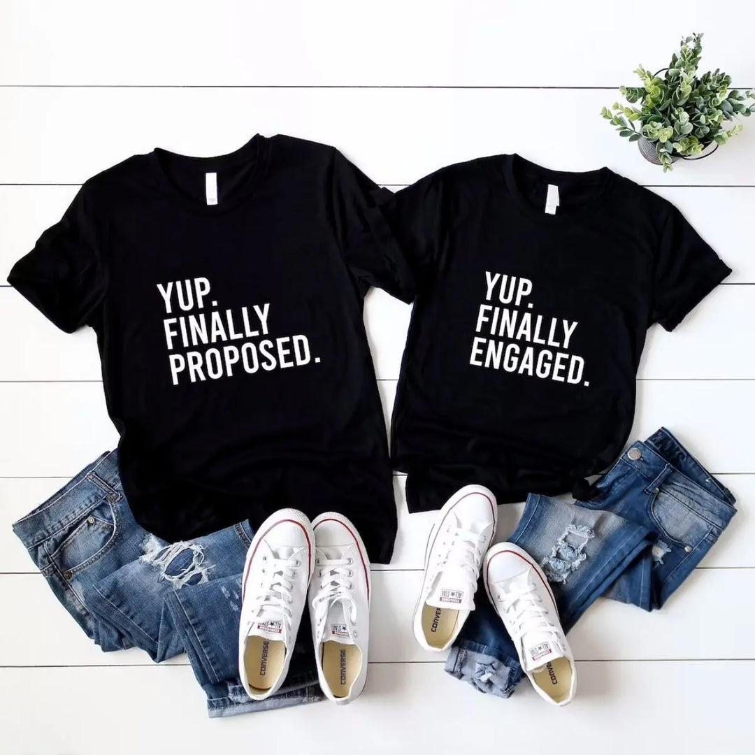Couples' matching black tees—his says 'Yup. Finally Proposed.' and hers reads 'Yup. Finally Engaged.' Bold white text for couples ready to celebrate the moment. Exclusively from CouplesChoices.com.