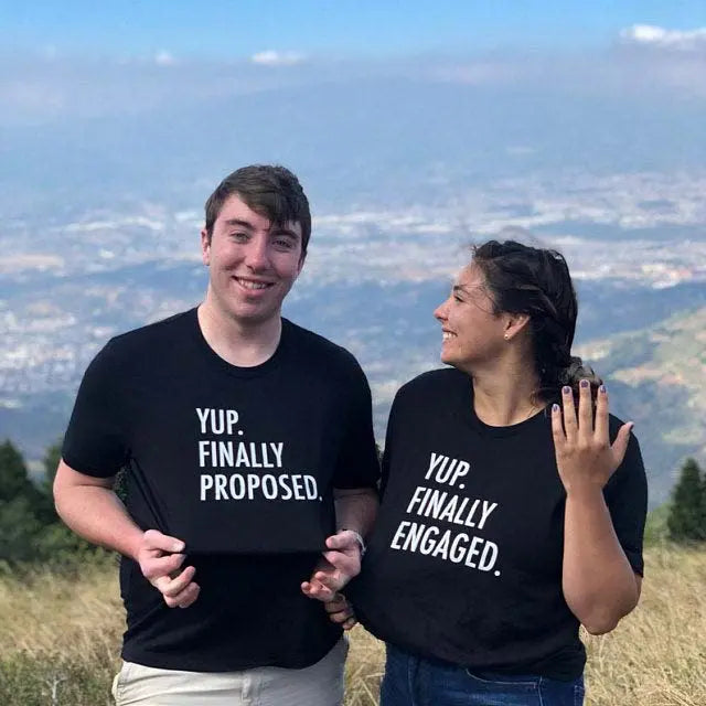 Couples' matching black tees—his says 'Yup. Finally Proposed.' and hers reads 'Yup. Finally Engaged.' Bold white text for couples ready to celebrate the moment. Exclusively from CouplesChoices.com.