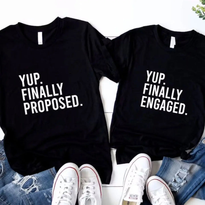 Couples' matching black tees—his says 'Yup. Finally Proposed.' and hers reads 'Yup. Finally Engaged.' Bold white text for couples ready to celebrate the moment. Exclusively from CouplesChoices.com.