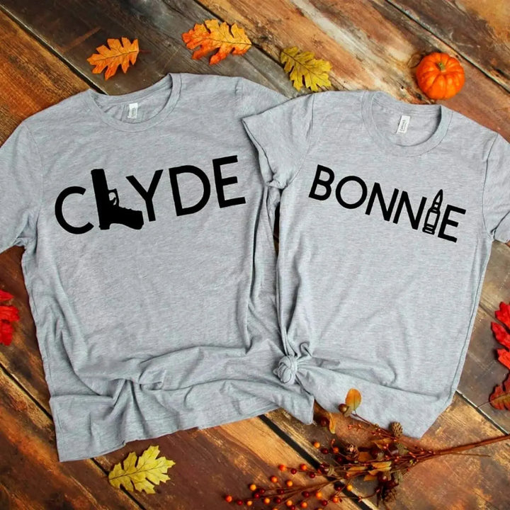 Matching Bonnie & Clyde couple shirts in soft heather grey, featuring bold black text with a gun and bullet design. Perfect for ride-or-die couples who love bold style and iconic vibes. Exclusively from CouplesChoices.com.