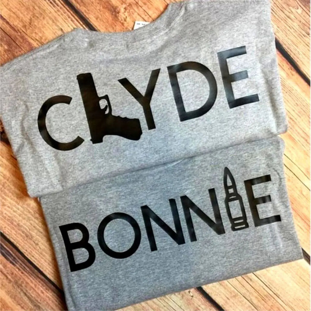 Matching Bonnie & Clyde couple shirts in soft heather grey, featuring bold black text with a gun and bullet design. Perfect for ride-or-die couples who love bold style and iconic vibes. Exclusively from CouplesChoices.com.