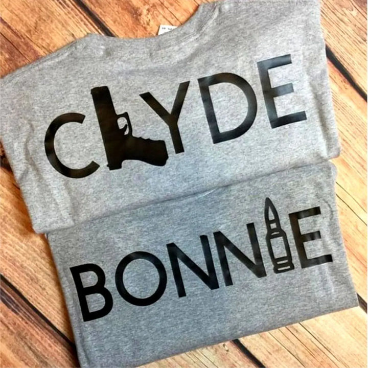 Matching Bonnie & Clyde couple shirts in soft heather grey, featuring bold black text with a gun and bullet design. Perfect for ride-or-die couples who love bold style and iconic vibes. Exclusively from CouplesChoices.com.