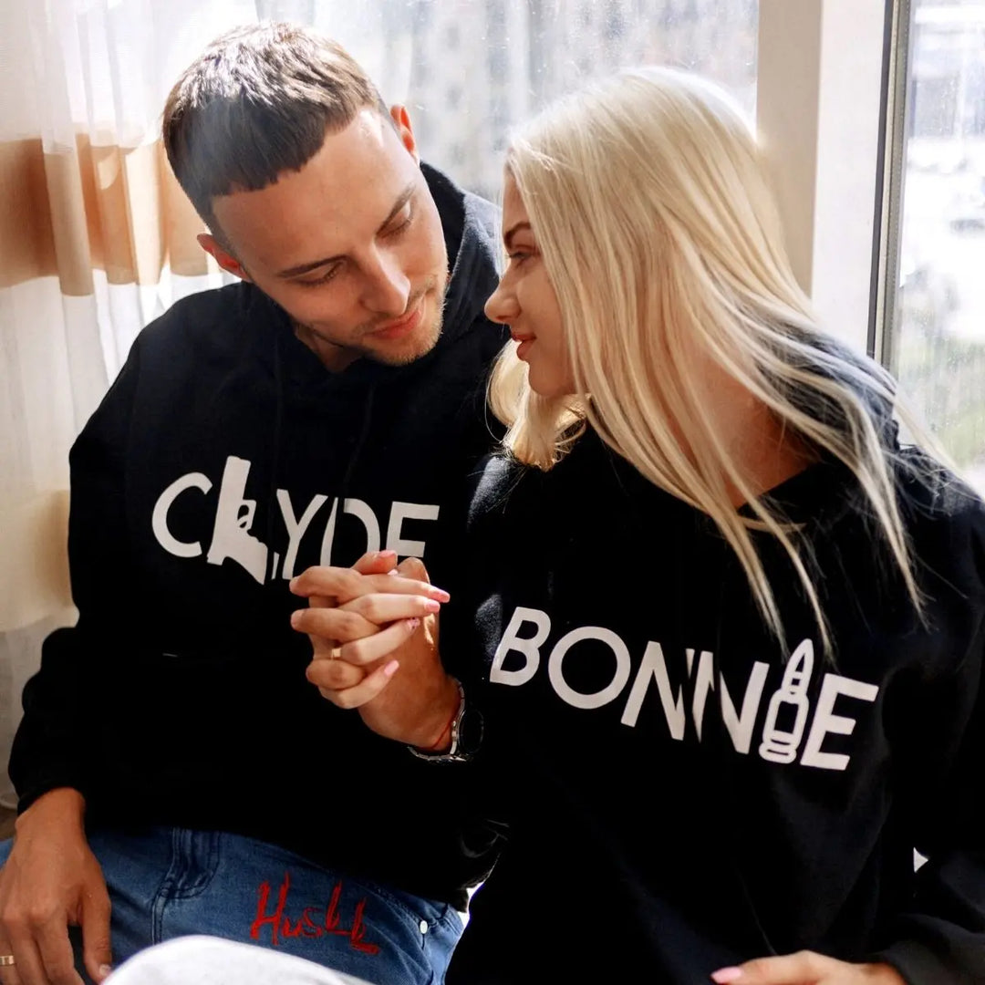 Matching black couple hoodies from CouplesChoices.com with bold white text — “BONNIE” featuring a bullet graphic and “CLYDE” with a gun silhouette. A daring and stylish set for couples who embrace loyalty, edge, and legendary love.