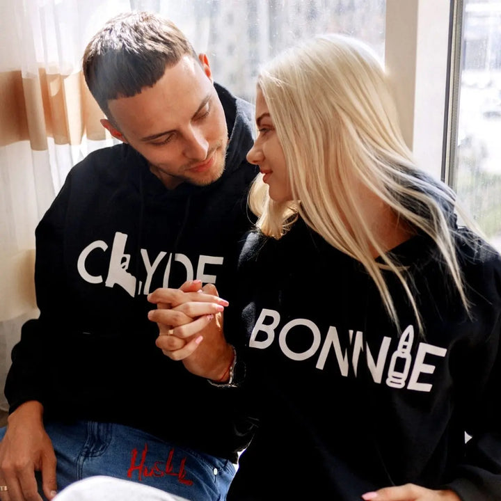 Matching black couple hoodies from CouplesChoices.com with bold white text — “BONNIE” featuring a bullet graphic and “CLYDE” with a gun silhouette. A daring and stylish set for couples who embrace loyalty, edge, and legendary love.