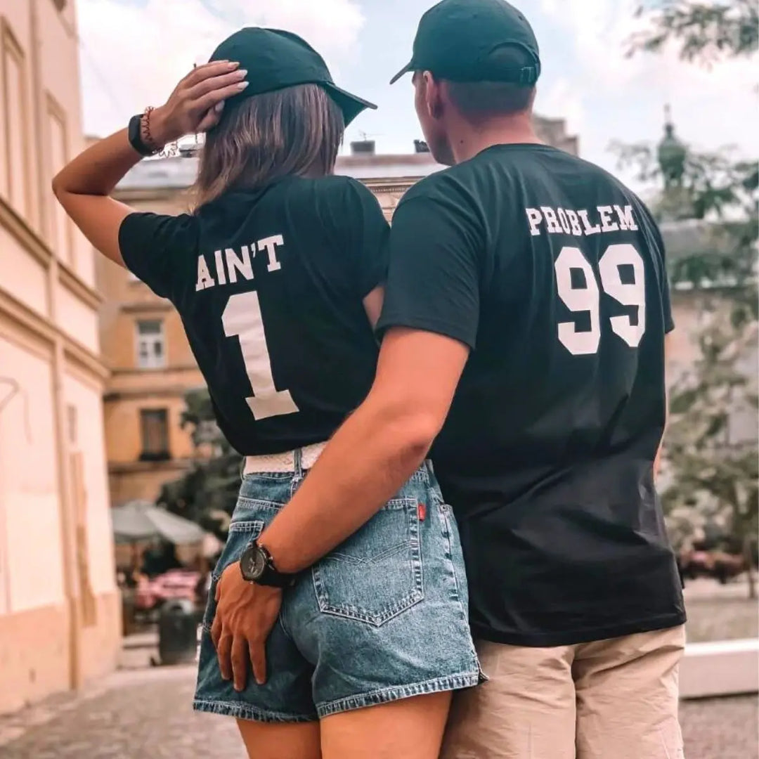 Matching black couple shirts—his reads '99 Problems' and hers answers 'Ain’t 1' in varsity-style white print. A playful nod to love and loyalty, exclusively from CouplesChoices.com.
